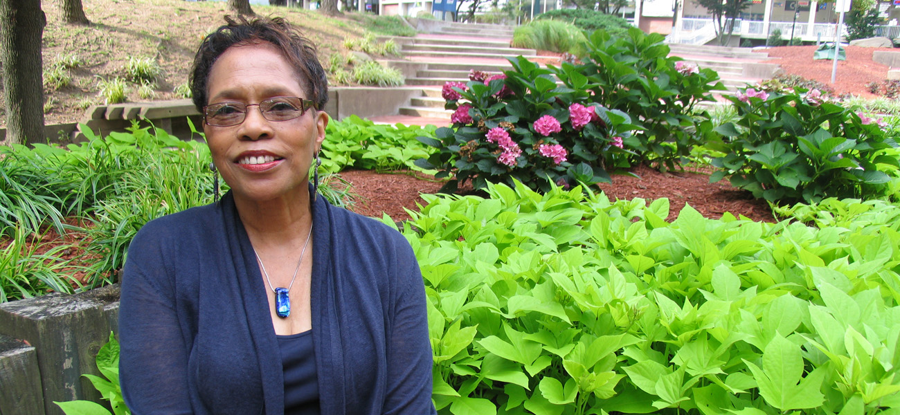 A smiling woman wearing glasses and a dark blue cardigan sits in front of lush green plants and blooming pink flowers in an outdoor garden setting.